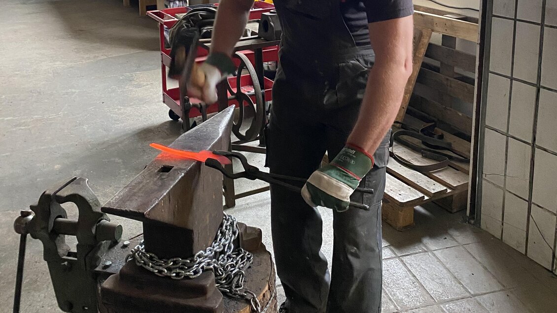 Ein Schmied bearbeitet heißes Metall an einem Amboss. Im Hintergrund sind Werkzeuge und Materialien sichtbar. | © Kurt Forstner