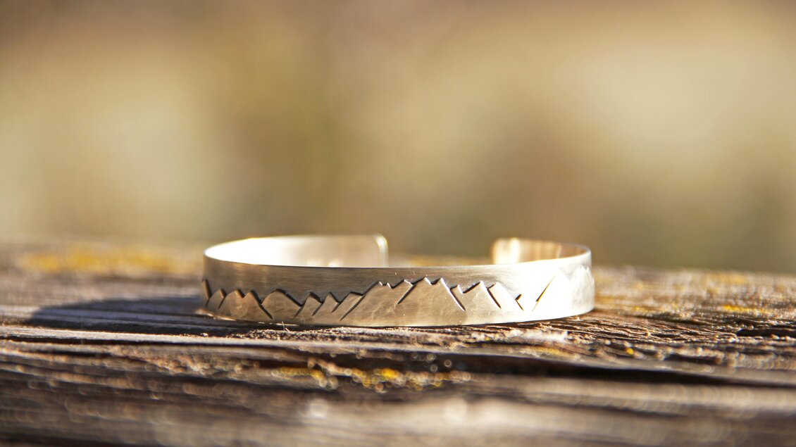 Ein schmaler Silberarmreif mit geometrischen Mustern liegt auf einem Holzbrett. Die natürliche Umgebung ist unscharf im Hintergrund zu sehen. | © Marlena Gerhart Schmuckgestalterin