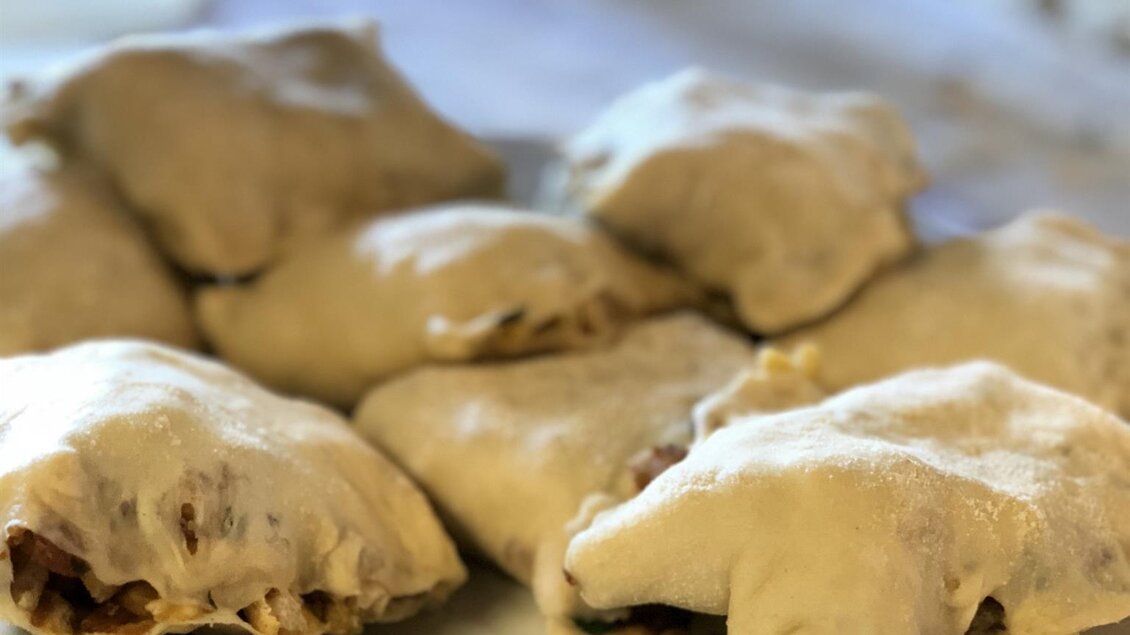 Ein Teller mit gefüllten Teigtaschen, die bereit zum Kochen sind. Im Hintergrund sind weitere Teigtaschen und eine Arbeitsfläche zu sehen. | © Michael Fischer