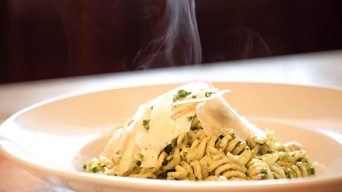 Ein Teller mit dampfender Pasta, garniert mit frischem Pesto und Parmesan. Die Nudelspiralen sind von grünen Kräutern umgeben. | © Corinna Harrer Fotografie