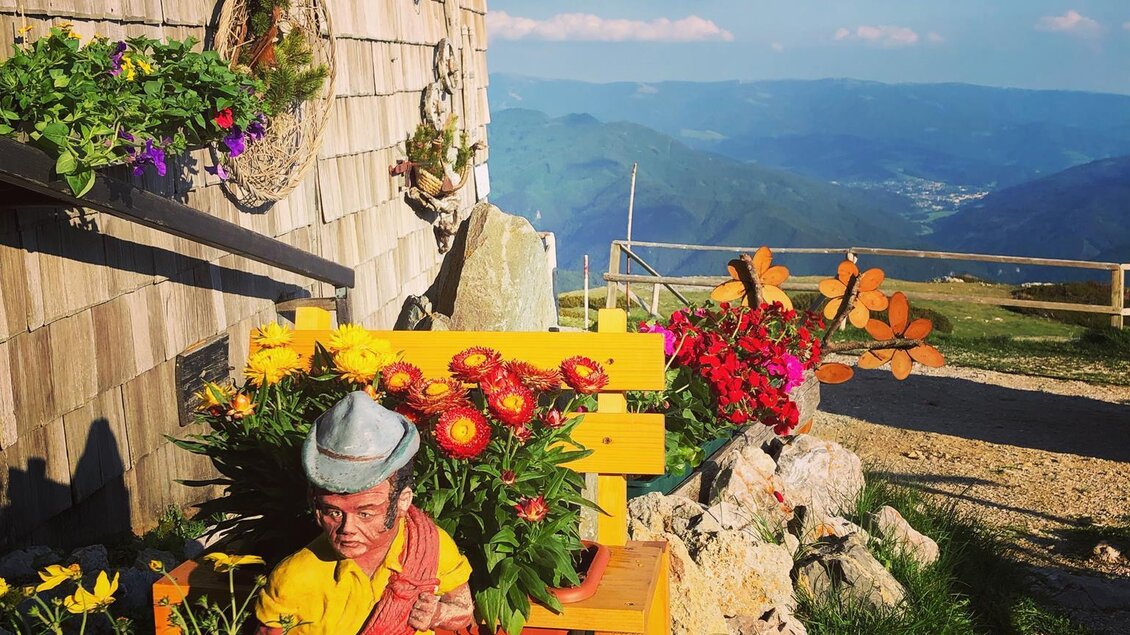 Eine bunte Figur sitzt auf einer Bank inmitten von Blumen. Im Hintergrund sind Berge und ein klarer Himmel zu sehen. | © Michael Fischer
