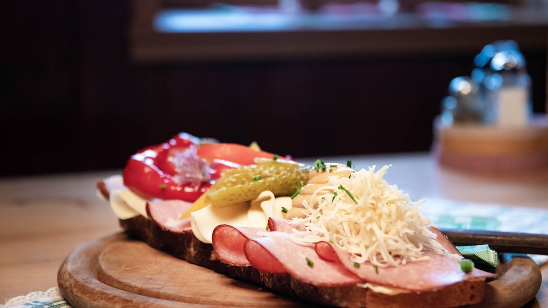 Ein belegtes Brot mit verschiedenen Wurstsorten, Käse und frischem Gemüse. Es liegt auf einem Holzbrett und ist appetitlich angerichtet. | © Corinna Harrer Fotografie