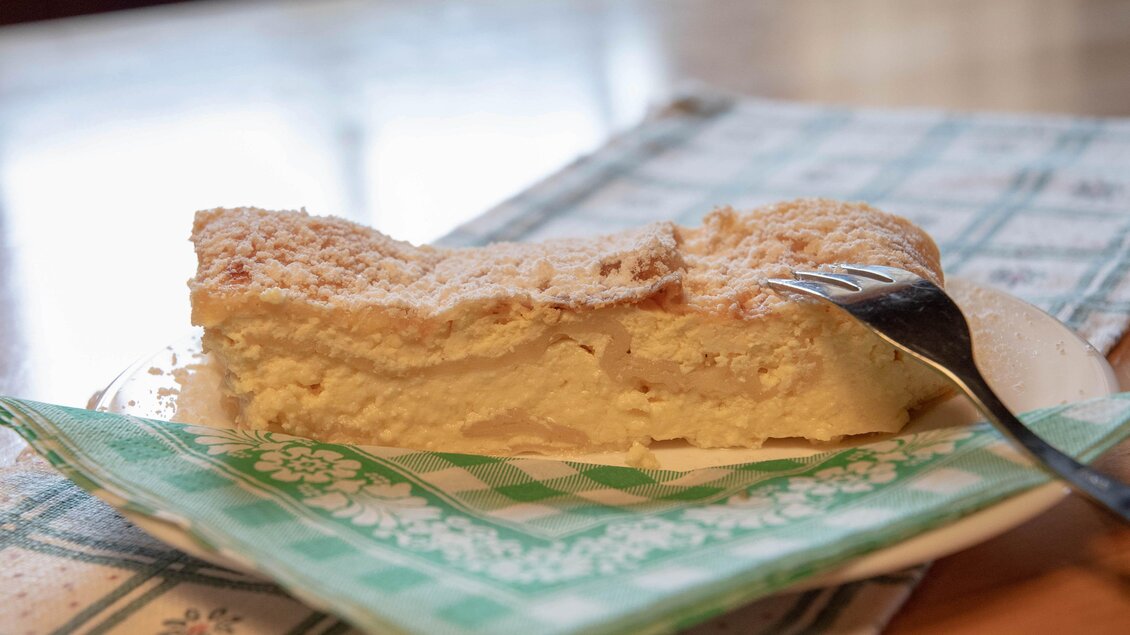 Ein Stück Käsekuchen auf einem Teller mit einer Gabel daneben. Der Kuchen liegt auf einer karierten Serviette. | © Corinna Harrer Fotografie