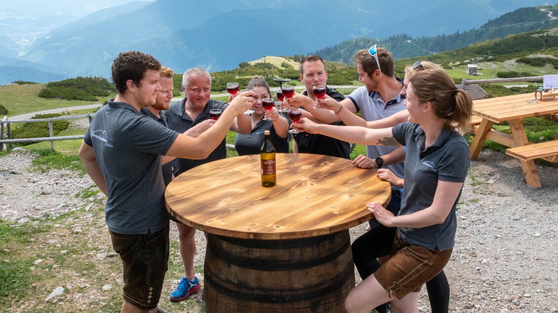 Eine Gruppe von Menschen hebt Gläser an einem Holztisch an einem malerischen Ort in den Bergen. Im Hintergrund sind sanfte Hügel und ein klarer Himmel zu sehen. | © Corinna Harrer Fotografie