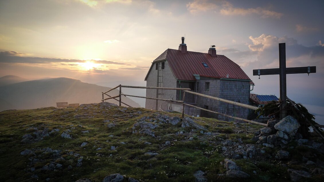 Eine malerische Berghütte mit rotem Dach steht auf einer Wiese. Im Hintergrund sieht man die untergehende Sonne und sanfte Hügel.