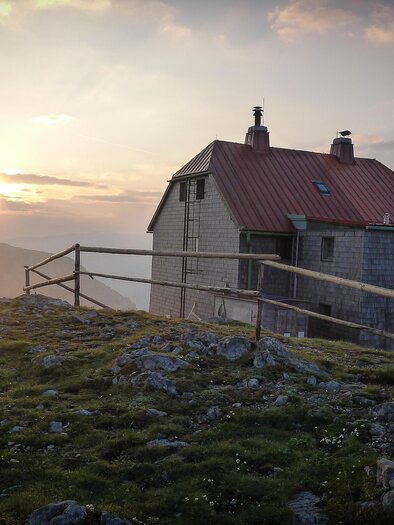 A picturesque mountain cottage with a red roof stands in a meadow. In the background, you can see the setting sun and gentle hills.