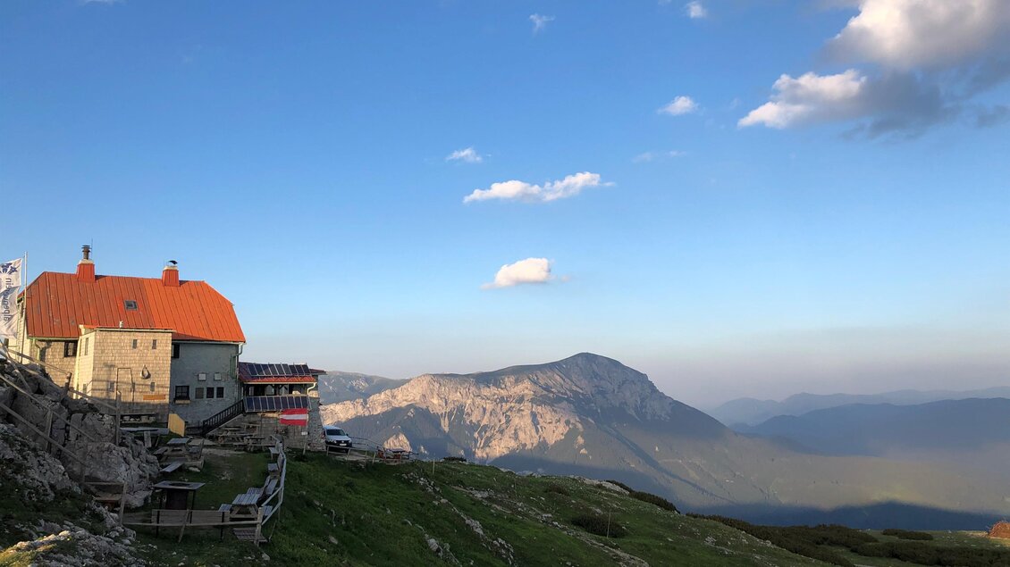 Ein malerisches Berghaus mit rotem Dach steht auf einer Wiese. Im Hintergrund sind majestätische Berge und ein blauer Himmel zu sehen.