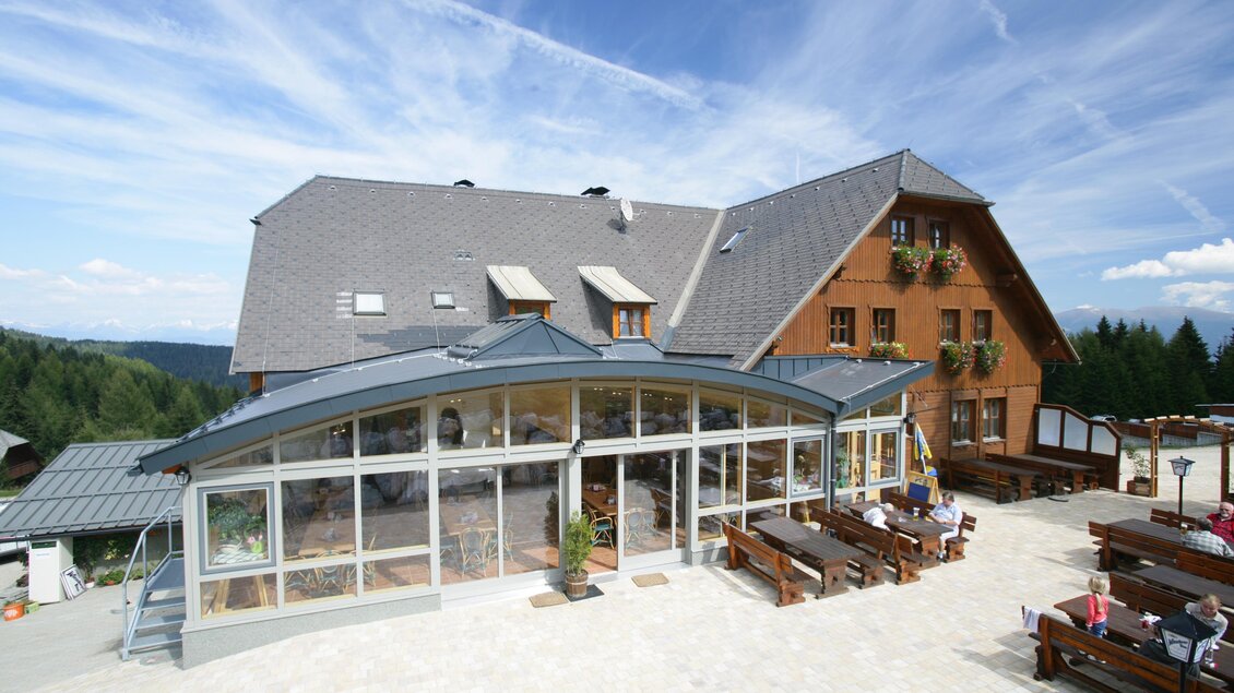 Ein einladendes Gasthaus mit einer großen Terrasse und gemütlichen Sitzgelegenheiten. Die Architektur verbindet Holzdesign mit modernen Elementen unter einem klaren blauen Himmel. | © Almidylle Sabathy