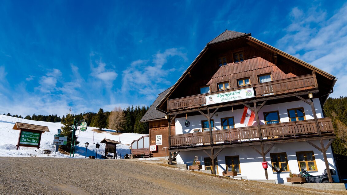 Ein traditionelles Berggasthaus mit Holzfassade und Balkon. Der Himmel ist blau und es liegt Schnee in der Umgebung. | © Almidylle Sabathy