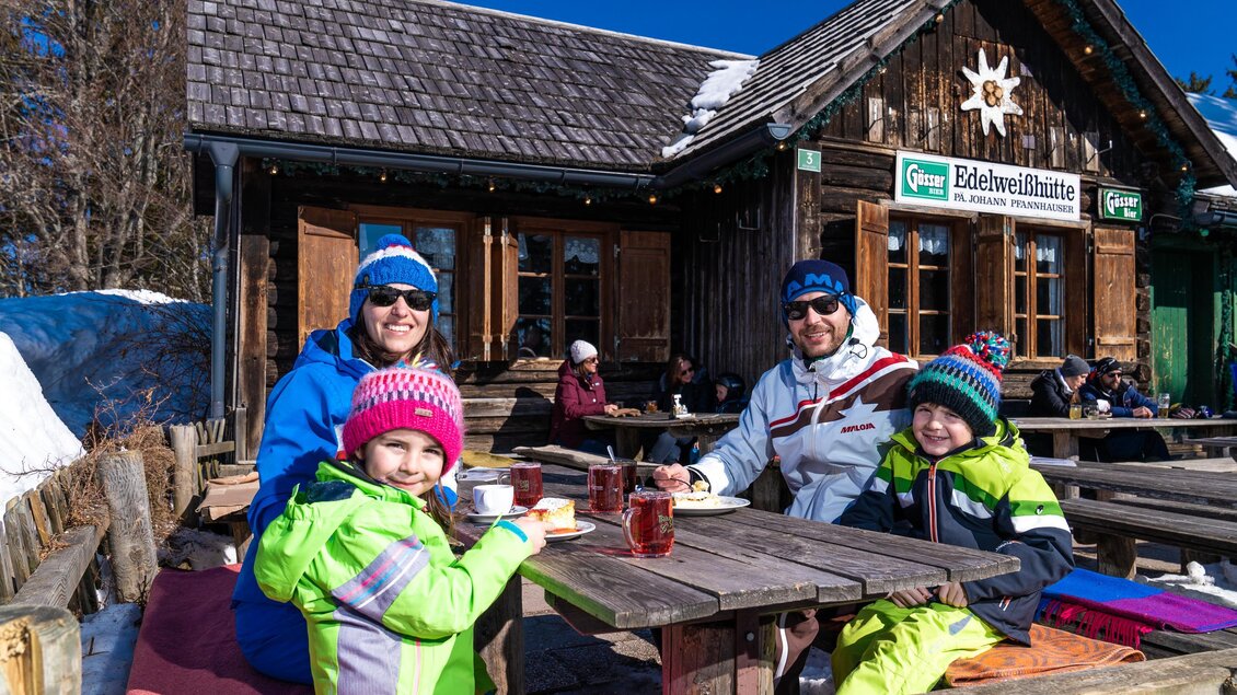 Eine Familie sitzt gemütlich im Freien an einem Tisch vor einer Berghütte. Es ist sonnig, und die Kinder tragen bunte Winterkleidung. | © Mariazeller Bürgeralpe | Rudy Dellinger