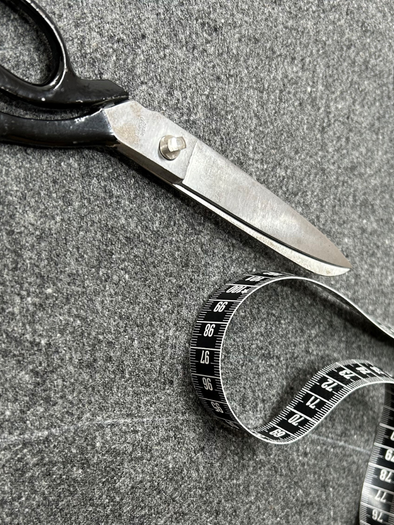 Eine Schere liegt auf einem grauen Untergrund neben einem Maßband. Das Maßband ist teilweise aufgerollt und zeigt Zahlen. | © Maria Greindl