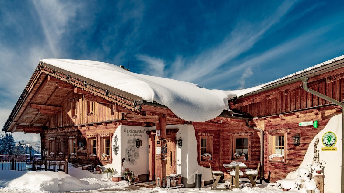 Ein traditionelles Holzhaus mit schneebedecktem Dach im Winter. Der blaue Himmel und die verschneite Umgebung schaffen eine malerische Atmosphäre. | © Christine Höflehner