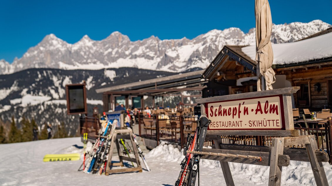 Eine Ski-Hütte namens "Schnepf’n-Alm" liegt in den Bergen mit schneebedeckten Landschaften. Im Vordergrund stehen Skier und die Sonne scheint auf den klaren blauen Himmel. | © Christine Höflehner