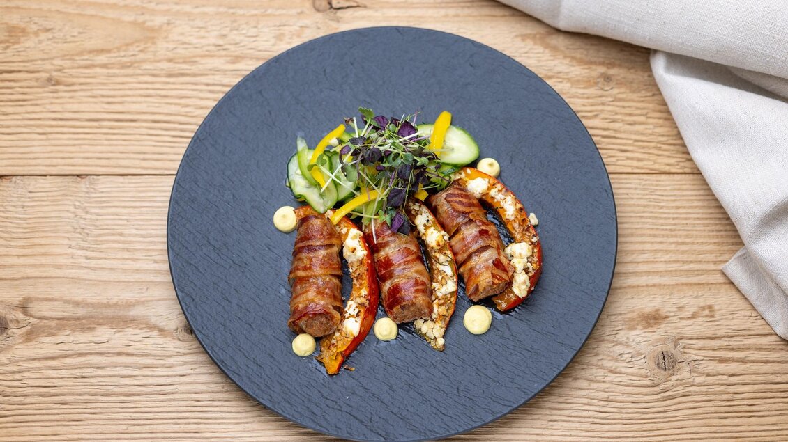 Ein Teller mit gefüllten Paprikaschoten, umwickelt mit Speck. Daneben frisches Gemüse und Saucen. | © Martin Huber