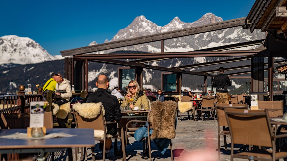 Eine sonnige Terrasse in den Bergen mit Gästen, die speisen und die Aussicht genießen. Im Hintergrund sind schneebedeckte Gipfel sichtbar. | © Christine Höflehner