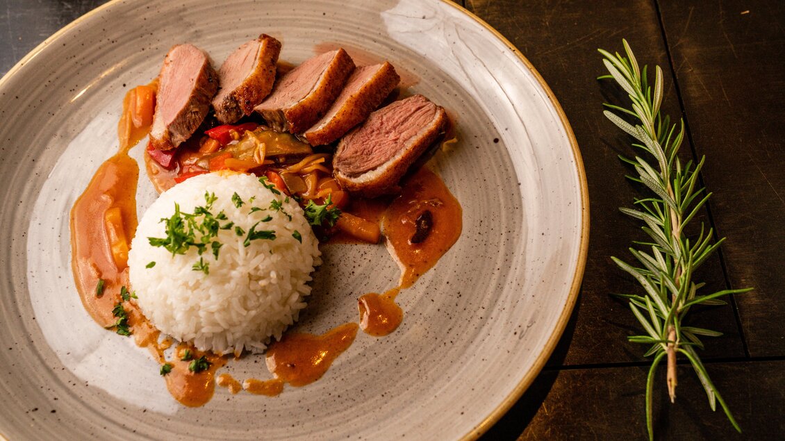 Ein Teller mit gebratenem Fleisch, begleitet von weißem Reis und buntem Gemüse in einer Sauce. Neben dem Teller liegt ein Zweig Rosmarin. | © Christine Höflehner