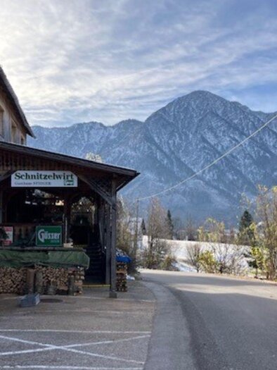 Ein gemütliches Gebäude mit einem Bistro-Schild steht an einer Straße. Im Hintergrund erheben sich schneebedeckte Berge unter einem klaren Himmel. | © TVB_Ausseerland Salzkammergut_Heike Temmel