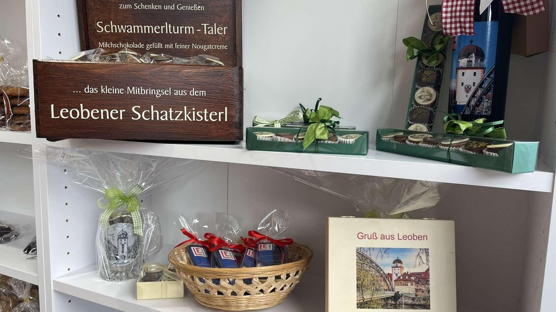 Ein Regal mit verschiedenen regionalen Produkten aus Leoben, einschließlich Geschenkartikeln und einer kleinen Schautafel. Darunter sind auch verpackte Süßigkeiten und ein Buch über Leoben zu sehen. | © TV Erzberg Leoben