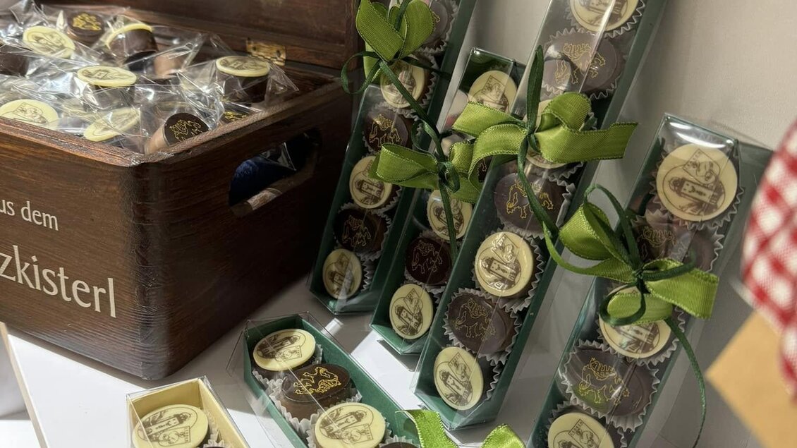 Eine schöne Geschenkverpackung mit Schokoladenstücken, die mit Bändern dekoriert sind. Im Hintergrund steht eine Holzkiste mit dem Aufdruck "Schwammerlturm-Taler". | © Sterner