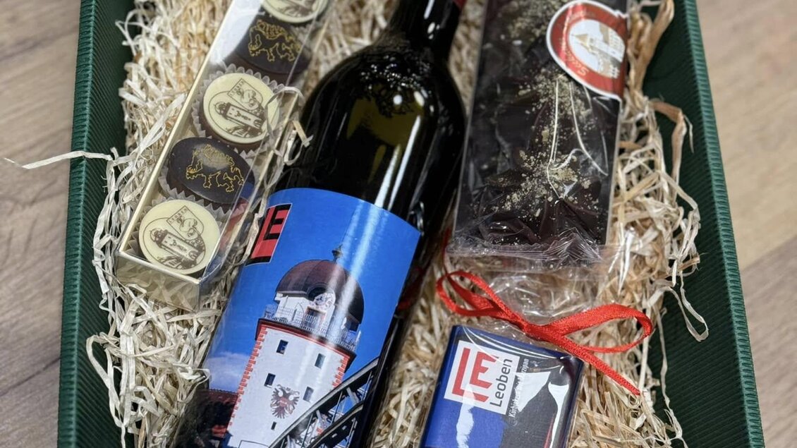 Ein Geschenkkorb mit einer Flasche Wein, Schokolade und einer kleinen Delikatesse. Alles ist kunstvoll in grünem Geschenkpapier arrangiert. | © Sterner