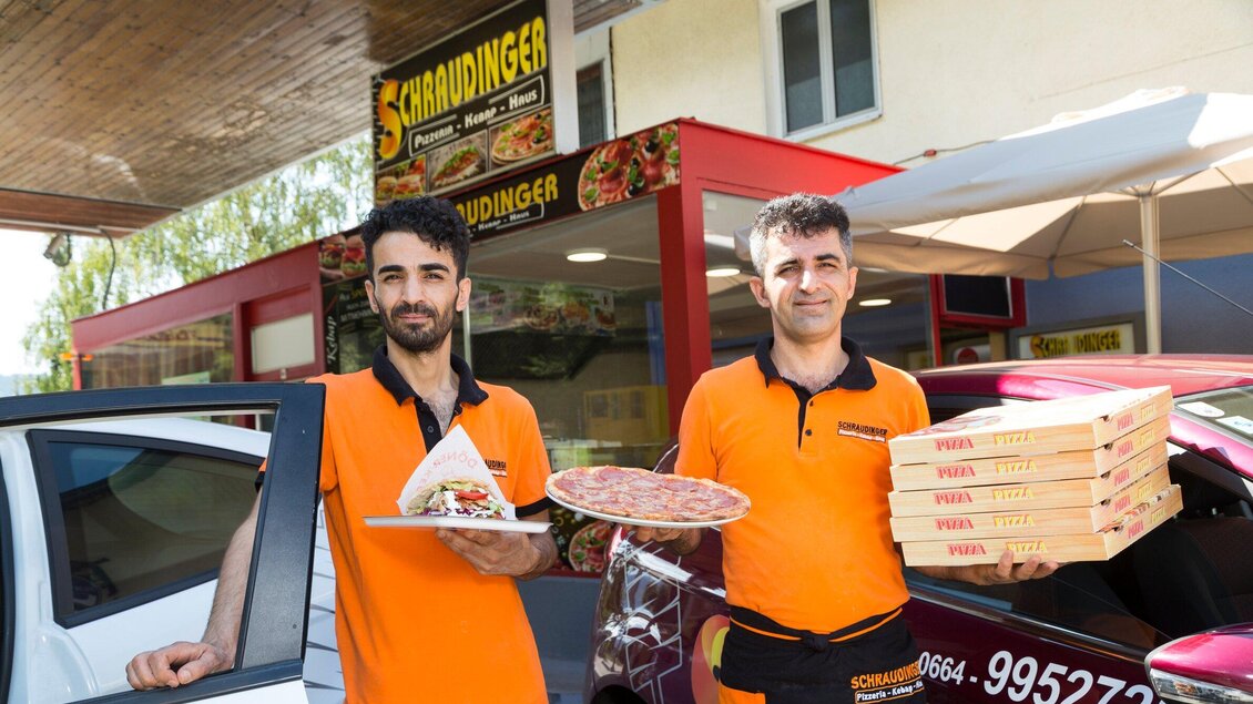 Zwei Männer in orangefarbenen T-Shirts stehen vor einem Restaurant und halten Essen in den Händen. Im Hintergrund ist das Restaurant mit einem großen Schild sichtbar. | © TV Region Graz - René Vidalli