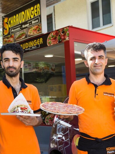 Zwei Männer in orangefarbenen T-Shirts stehen vor einem Restaurant und halten Essen in den Händen. Im Hintergrund ist das Restaurant mit einem großen Schild sichtbar. | © TV Region Graz - René Vidalli