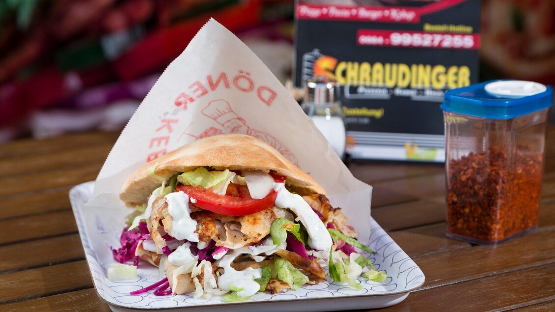 Ein leckerer Döner in einem Fladenbrot, gefüllt mit frischem Gemüse, Joghurtsoße und scharfer Sauce. Im Hintergrund sind Gewürze und eine Tafel sichtbar. | © TV Region Graz - René Vidalli