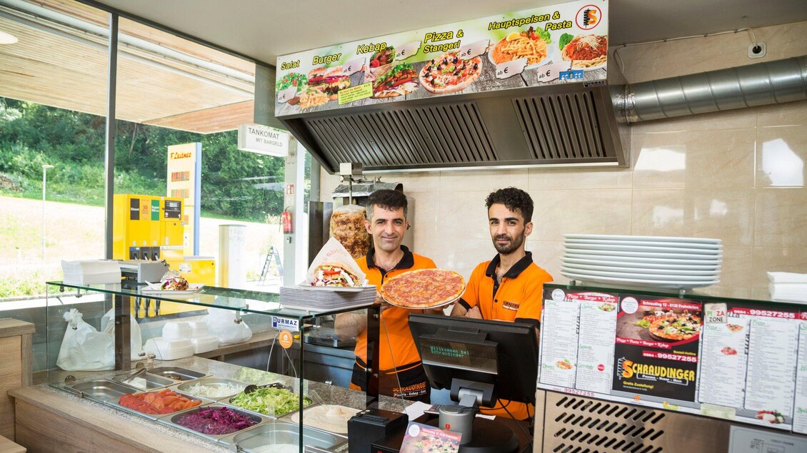 Ein Imbiss mit freundlichen Mitarbeitern, die frisch zubereitete Speisen präsentieren. Im Hintergrund sind verschiedene Zutaten und eine Speisekarte sichtbar. | © TV Region Graz - René Vidalli
