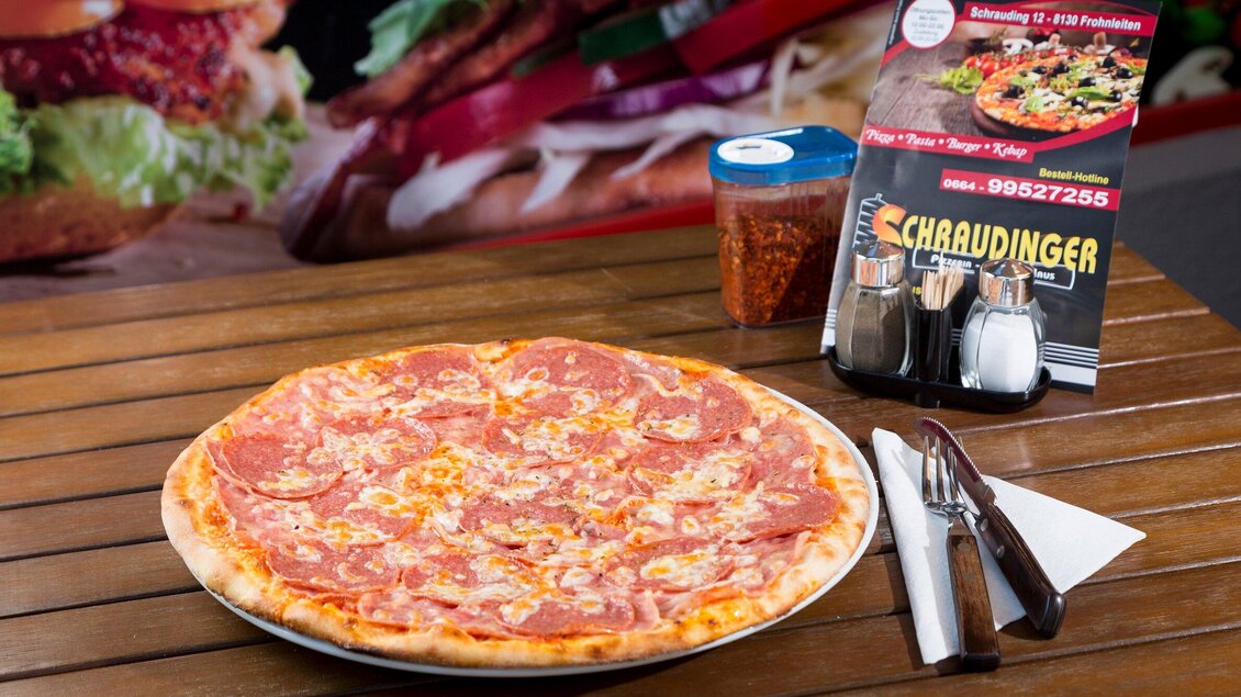 Eine große Pizza mit Salami und Käse auf einem Holztisch. Daneben steht ein Menü und ein Glas mit Gewürzen. | © TV Region Graz - René Vidalli