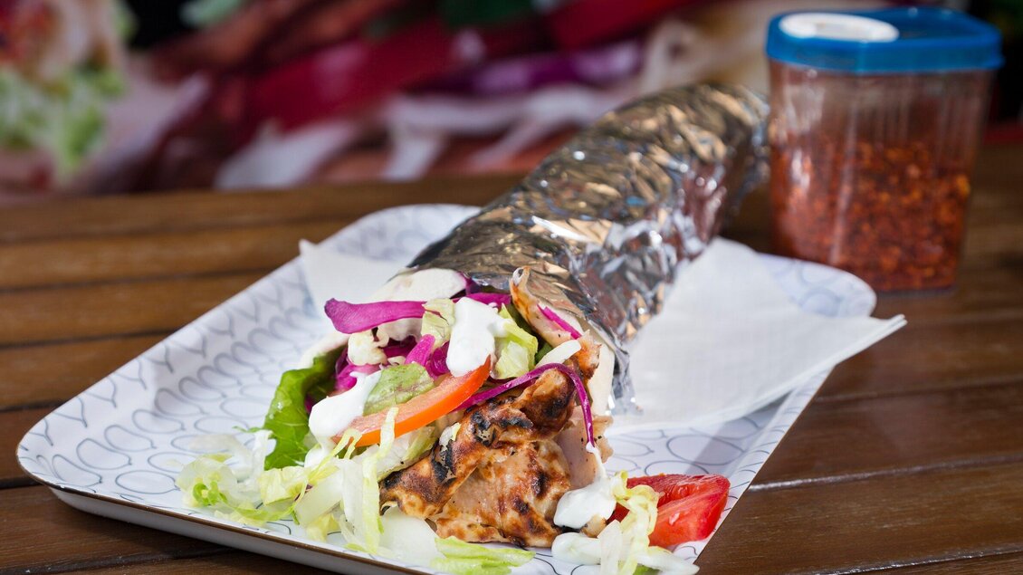 Ein leckerer Burrito, in Folie gewickelt, liegt auf einem Teller. Neben dem Burrito sieht man frisches Gemüse und eine Schale mit Chili. | © TV Region Graz - René Vidalli