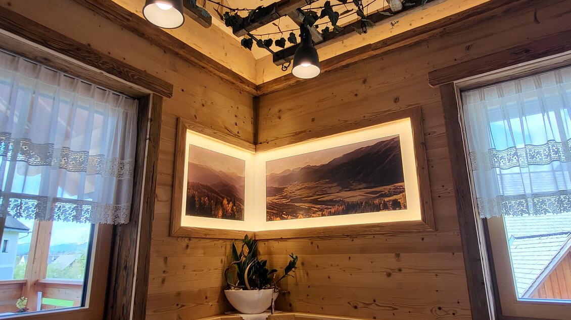 Eine gemütliche Holzwand mit Fenstern und einem beleuchteten Bild. Grüne Pflanzen und eine einladende Sitzgelegenheit vervollständigen die Atmosphäre. | © TV Hochsteiermark
