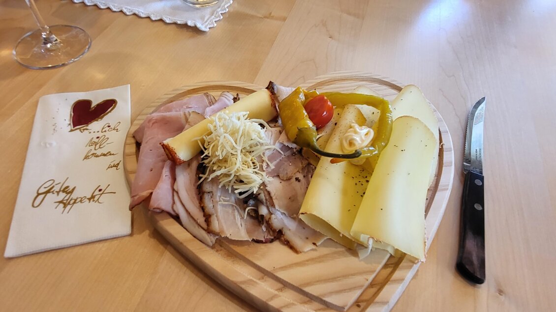 Eine herzförmige Holzplatte mit verschiedenen Wurst- und Käsesorten, garniert mit Gemüse. Daneben liegt eine Serviette mit einem herzlichen Gruß. | © TV Hochsteiermark
