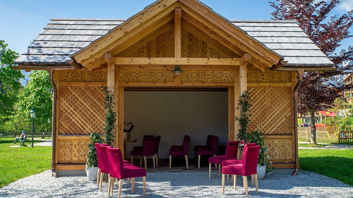 Ein gemütliches Holzhaus mit einem offenen Eingangsbereich. Vor dem Haus stehen rote Stühle auf einem Pflasterboden. | © Lebensbilder_Christine-Struz