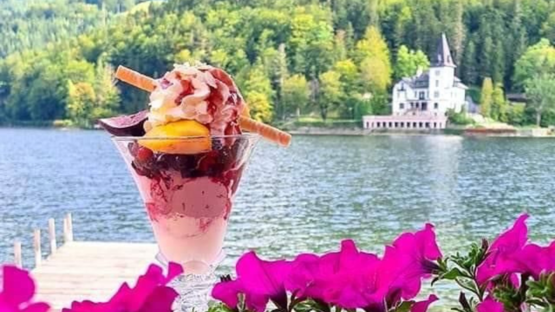 Ein köstliches Eis in einem Glas mit bunten Früchten und Waffeln. Im Hintergrund sieht man einen malerischen See und ein schönes Gebäude. | © Christoph Knappe