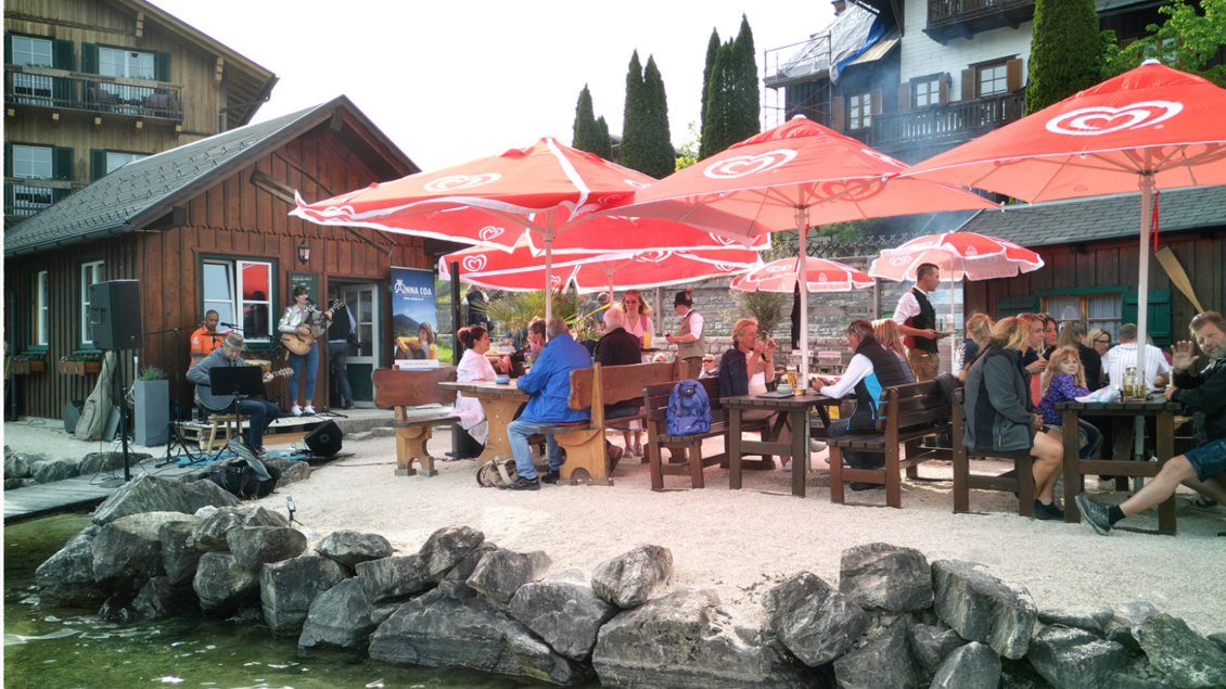 Ein gemütliches Restaurant am Wasser mit Tischen unter roten Sonnenschirmen. Gäste genießen das Essen und die Musik in einer schönen Umgebung. | © Christoph Knappe