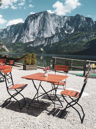 A beautiful outdoor area with wooden chairs and tables by the shore of a lake. In the background, tall mountains and a clear sky can be seen. | © Theresa Schwaiger