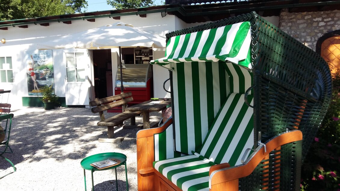 Ein Strandkorb mit grünen und weißen Streifen steht vor einem kleinen, einladenden Gebäude. Es gibt einige Sitzgelegenheiten im Freien und viel Tageslicht. | © TVB Ausseerland - Salzkammergut | Petra Kirchschla
