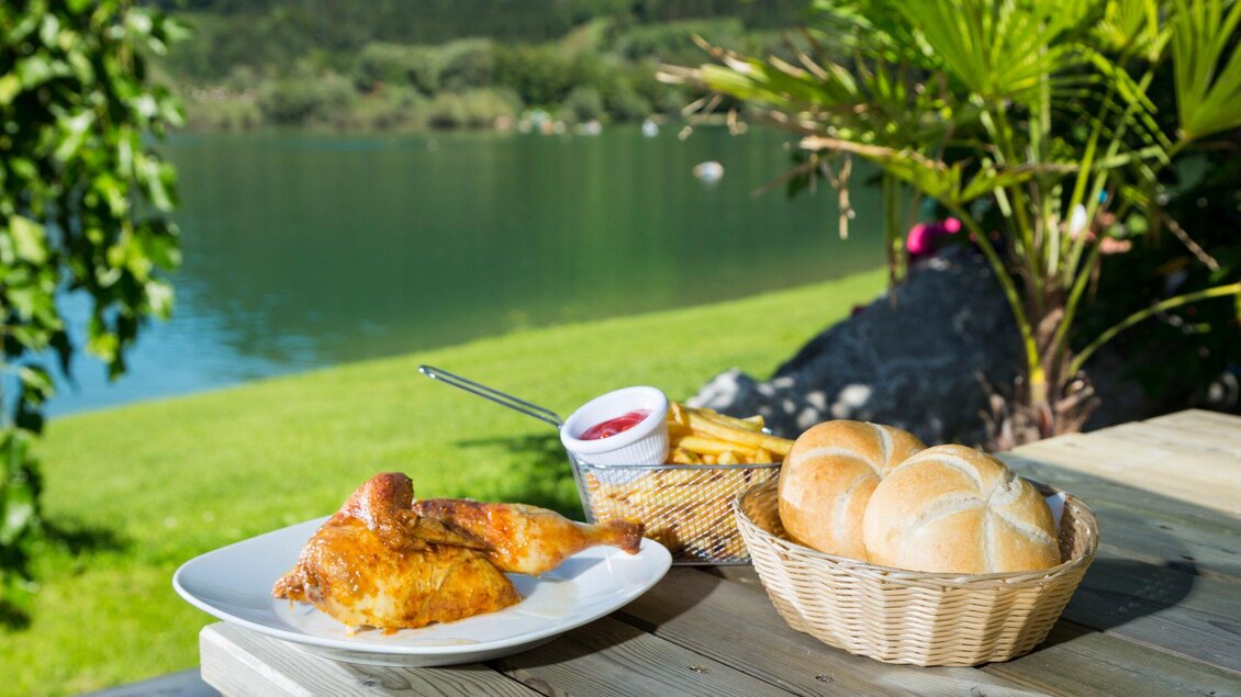 Ein Teller mit knusprigem Hähnchen, Pommes und Sauce steht auf einem Tisch. Im Hintergrund ist ein ruhiger See und grüne Wiesen zu sehen. | © TV Region Graz - René Vidalli