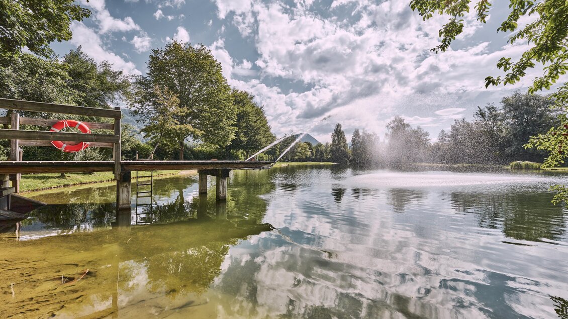 Ein ruhiger Teich mit klarer Wasseroberfläche und üppigem Grün. Im Hintergrund sprudelt ein Wasserstrahl und der Himmel ist teilweise bewölkt. | © Thomas Sattler