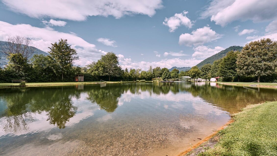 Ein ruhiger See umgeben von Bäumen und grünen Wiesen. Strahlend blauer Himmel mit wenigen Wolken. | © Thomas Sattler