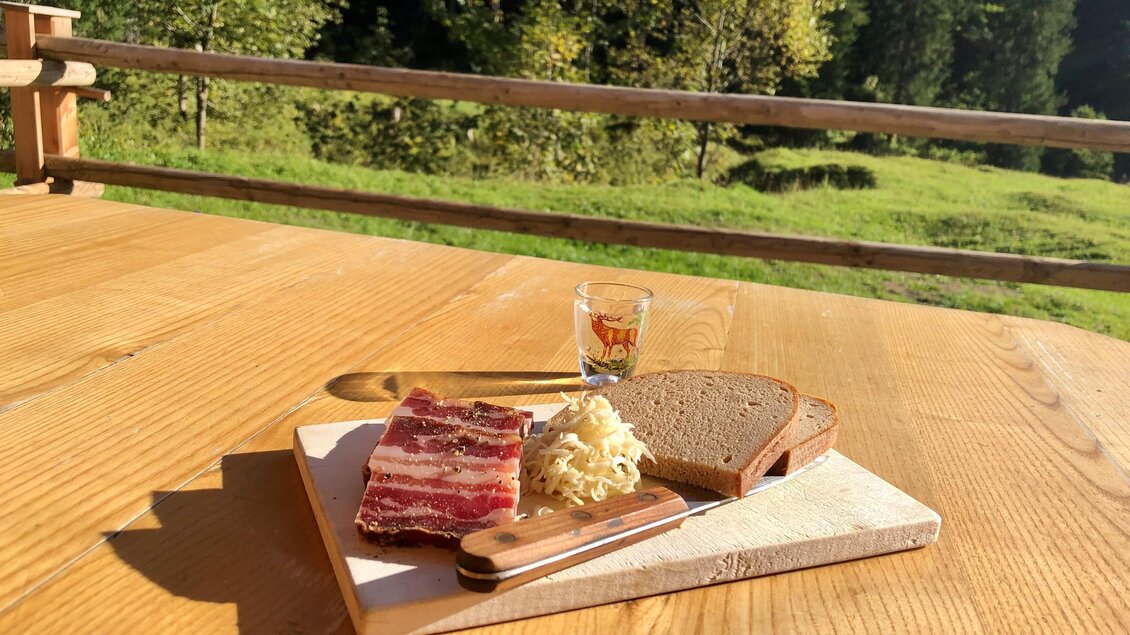 Ein rustikales Frühstücksbrett mit Brot, Speck und Käse, begleitet von einem Schnaps. Im Hintergrund ist eine grüne Landschaft zu sehen. | © Gertraud Kalss