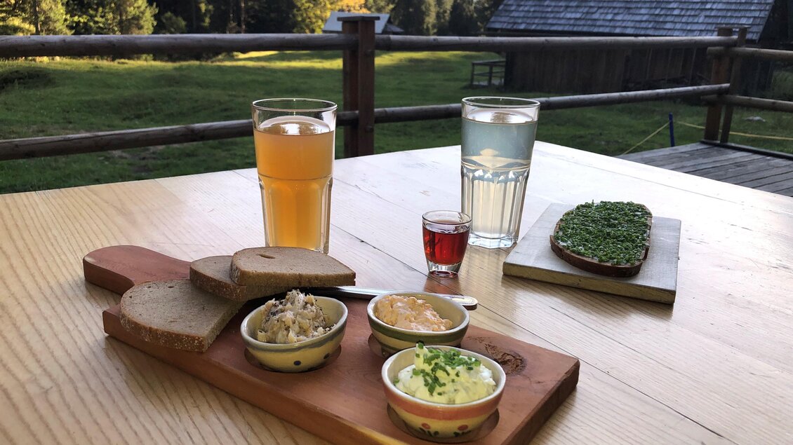 Ein rustikales Frühstücksbrett mit verschiedenen Aufstrichen, frischem Brot und Getränken. Im Hintergrund sind grüne Wiesen und Bäume zu sehen. | © Gertraud Kalss