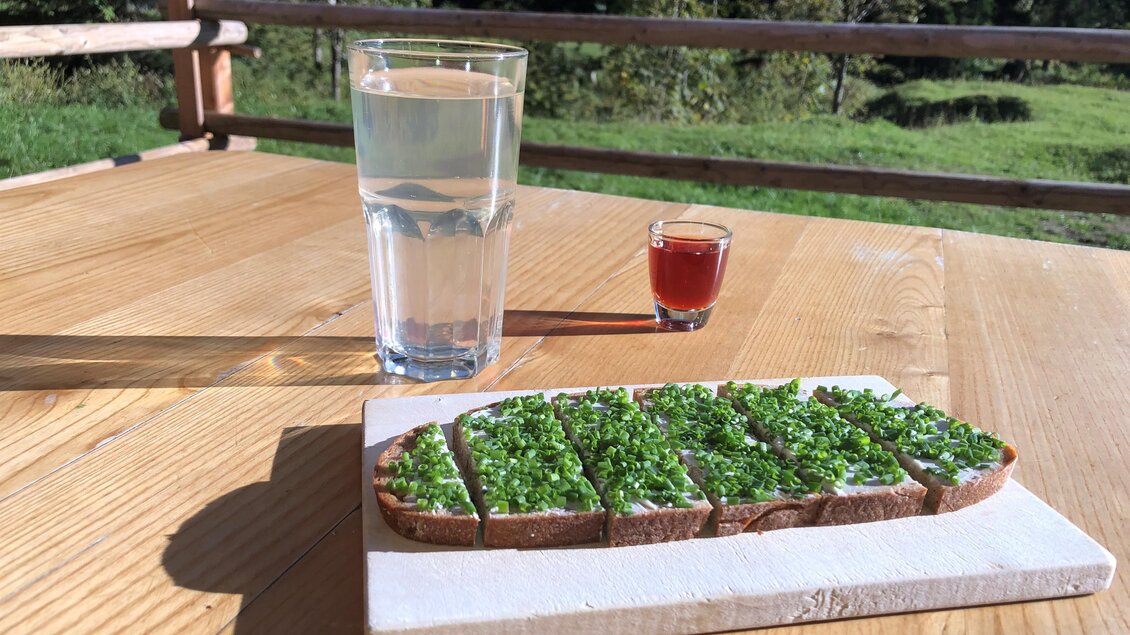Ein Holzbrett mit einer belegten Brotscheibe, die mit frischem Schnittlauch garniert ist. Daneben stehen ein Glas Wasser und ein kleiner Schnaps auf einem Tisch im Freien. | © Gertraud Kalss