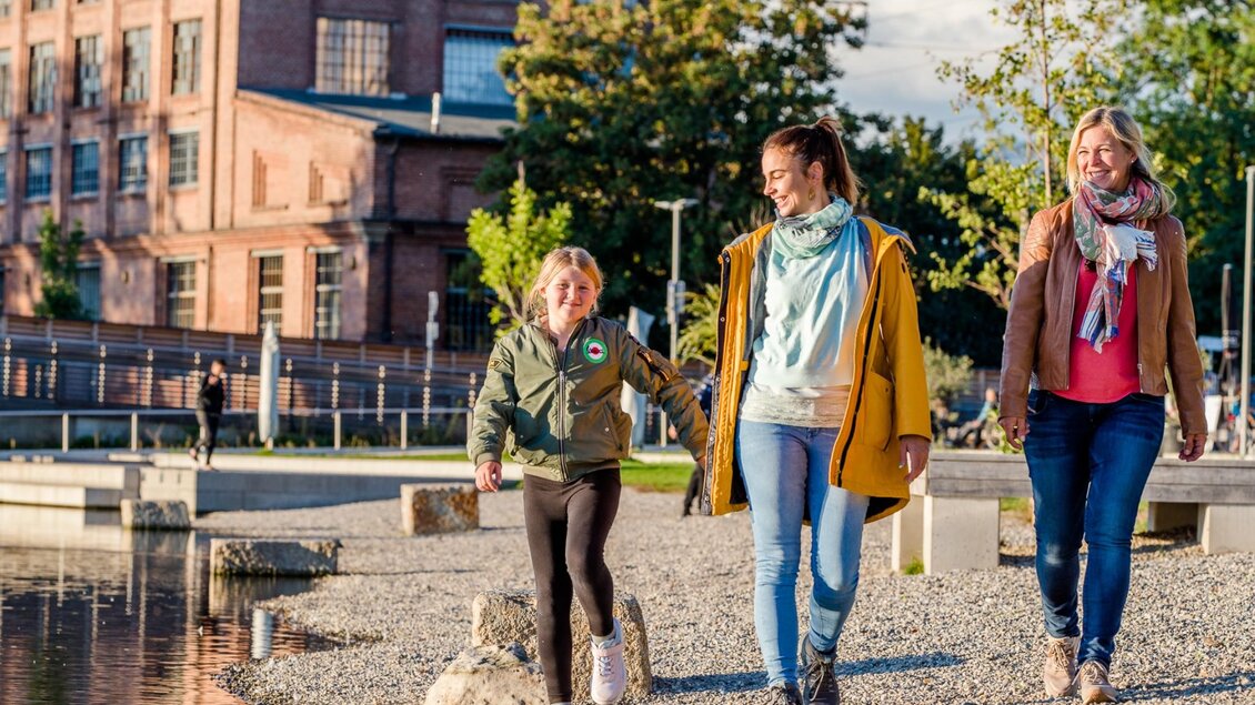 Drei Personen spazieren am Wasser entlang vor der roten Ziegelfassade der Seifenfabrik Graz. | © Graz Tourismus-Mias Photoart