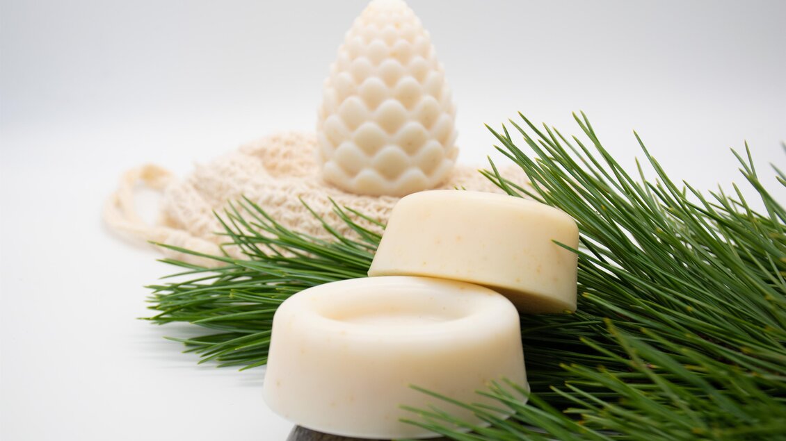 Eine Reihe von weißen Seifen liegt auf grünen Zweigen. Im Hintergrund steht eine kegelförmige Kerze. | © lela. Design Studio