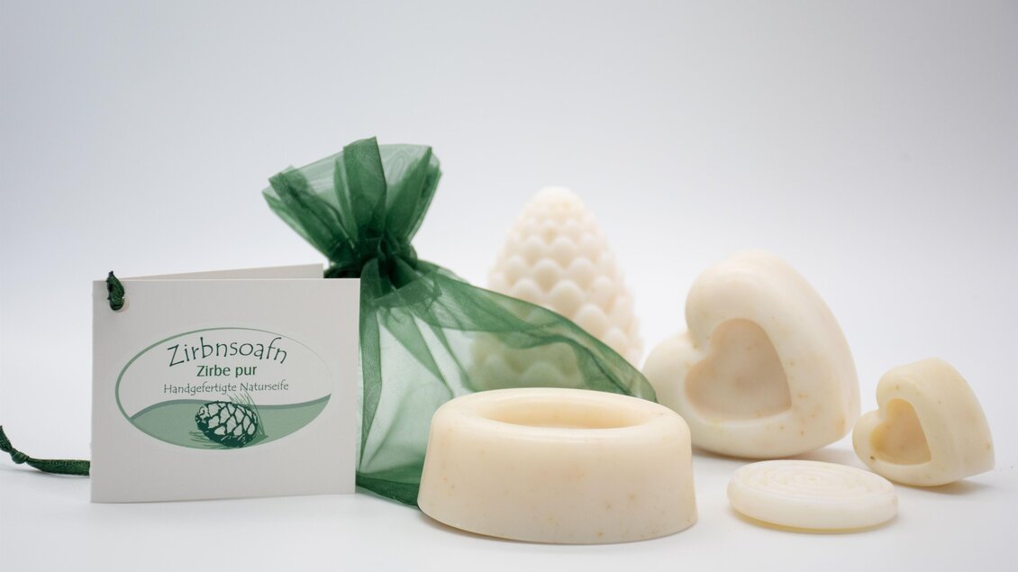 Eine Auswahl an handgemachten Seifen in verschiedenen Formen, darunter Herzen und eine Tannenzapfenform. Die Seifen sind in einem grünen Organza-Beutel verpackt, zusammen mit einem Etikett. | © lela. Design Studio