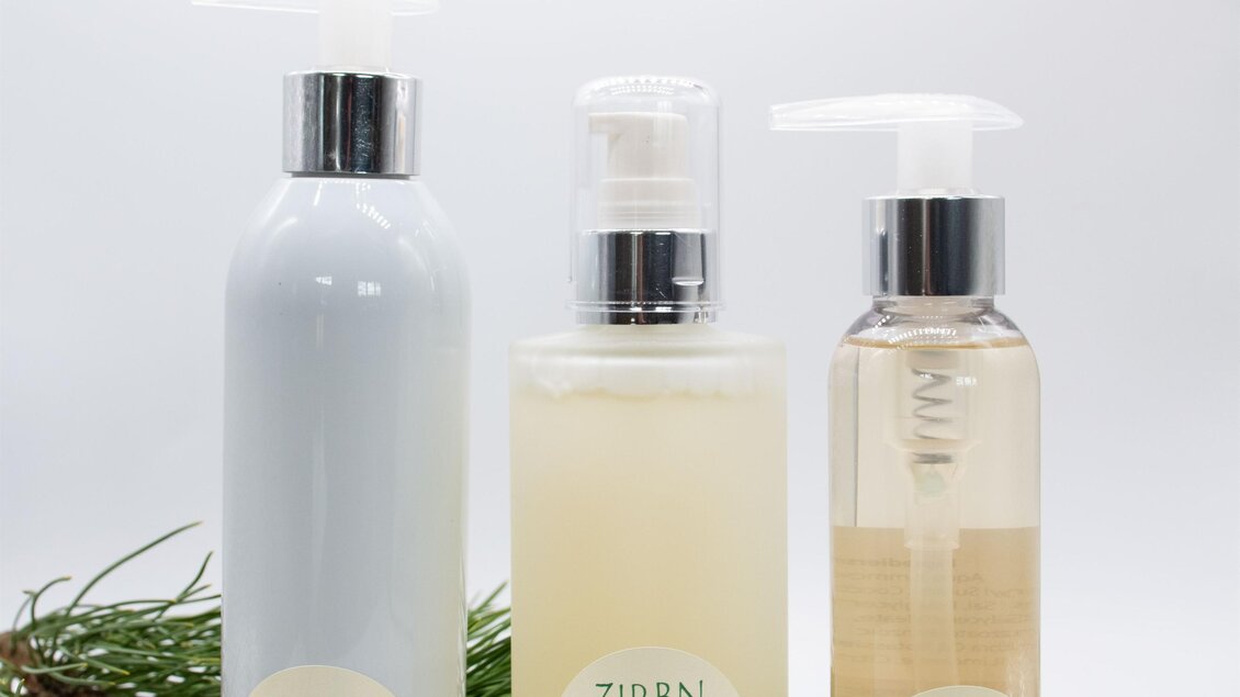Drei Flaschen mit Zirbenduschgel in unterschiedlichen Größen stehen vor einem weißen Hintergrund. Die Produkte sind von einem Zweig mit Tannennadeln umgeben. | © lela. Design Studio