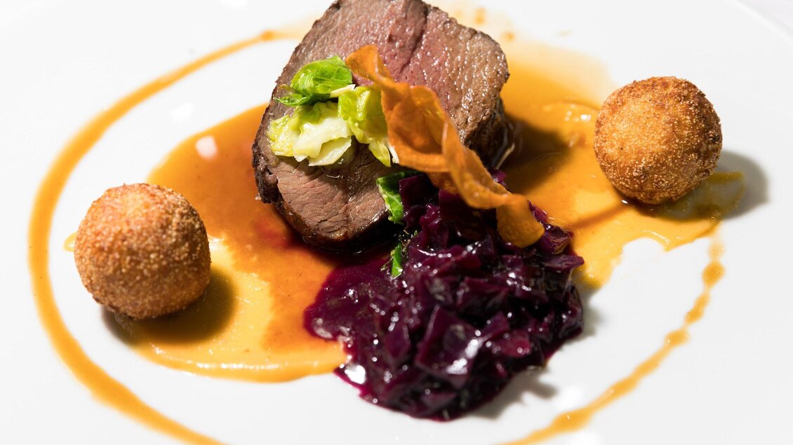 Ein köstliches Gericht mit zartem Fleisch, serviert mit Rotkohl und zwei knusprigen Bällchen. Die Sauce auf dem Teller rundet das Essen perfekt ab. | © TV Region Graz - Harry Schiffer