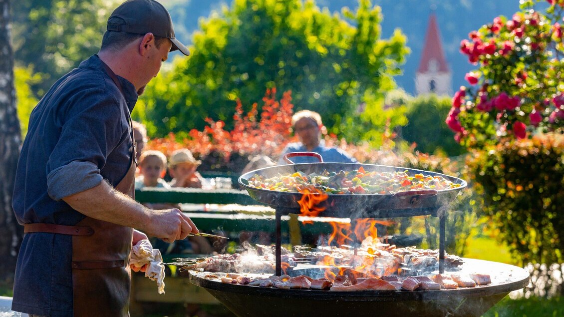 Ein Mann grillt im Freien mit einem großen Grill. Im Hintergrund sitzen Menschen und genießen das schöne Wetter. | © TV Region Graz - Harry Schiffer