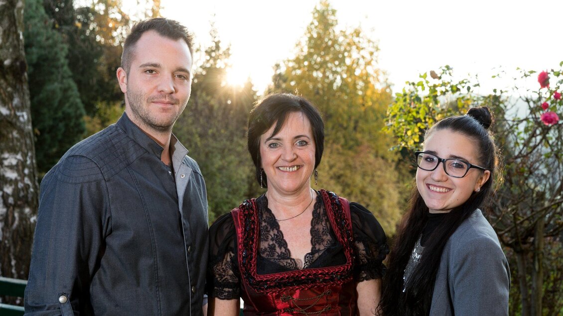 Drei Personen stehen lächelnd im Freien, umgeben von Bäumen und Blumen. Die Frau in der Mitte trägt ein traditionelles Kleid. | © TV Region Graz - Harry Schiffer